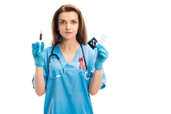 Atractivo médico con la cinta roja que sostiene la jeringa con sangre y condón con las píldoras aisladas en el concepto blanco, del día del sida del mundo — Stock Photo