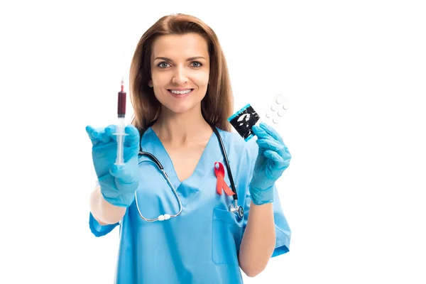 Atractivo médico sonriente con la cinta roja que sostiene la jeringa con sangre y condón con las píldoras aisladas en el concepto blanco, del día del SIDA del mundo — Stock Photo