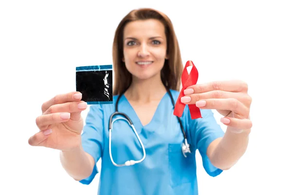 Smiling attractive doctor holding condom and red ribbon isolated on white, world aids day concept — Stock Photo