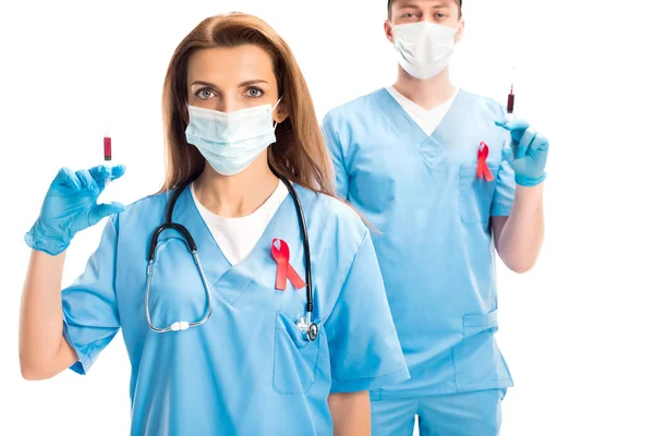 Médicos en máscaras médicas y cintas rojas que sostienen jeringas con sangre aislada en el concepto del día del sida blanco, mundial - foto de stock