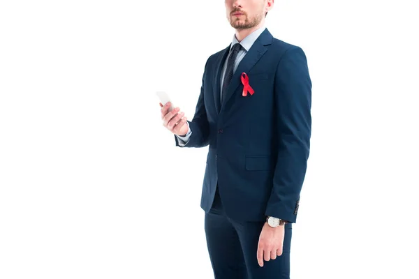 Imagen recortada de hombre de negocios con cinta roja en traje que sostiene el teléfono inteligente aislado en blanco, concepto del día del sida mundial - foto de stock
