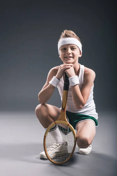 Улыбающийся мальчик в спортивной одежде с теннисной ракеткой на темном фоне — стоковое фото