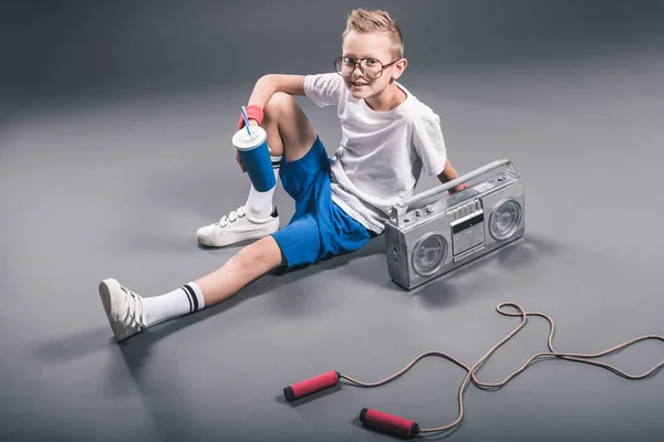 Vista de ángulo alto de niño en gafas con bebida gaseosa, boombox y cuerda de salto sobre fondo gris - foto de stock