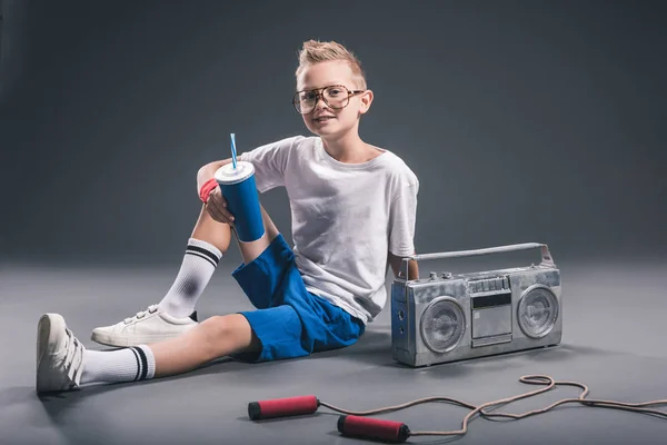 Niño en gafas con bebida gaseosa, boombox y cuerda de salto sobre fondo gris - foto de stock