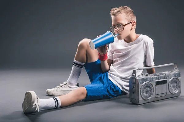 Stylish boy in eyeglasses drinking soda drink with boombox near by on grey background — Stock Photo
