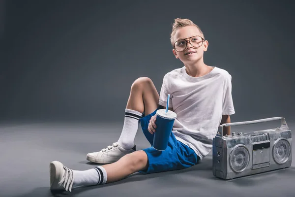 Menino elegante em óculos com bebida de refrigerante e boombox no fundo cinza — Fotografia de Stock