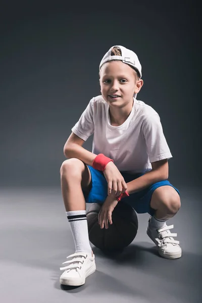 Stylish boy sitting on basketball ball on grey background — Stock Photo