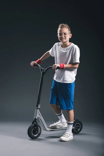 Smiling stylish boy in eyeglasses on scooter looking at camera on grey backdrop — Stock Photo