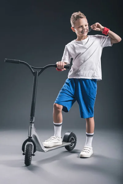 Stylish boy in eyeglasses with scooter looking away on grey backdrop — Stock Photo