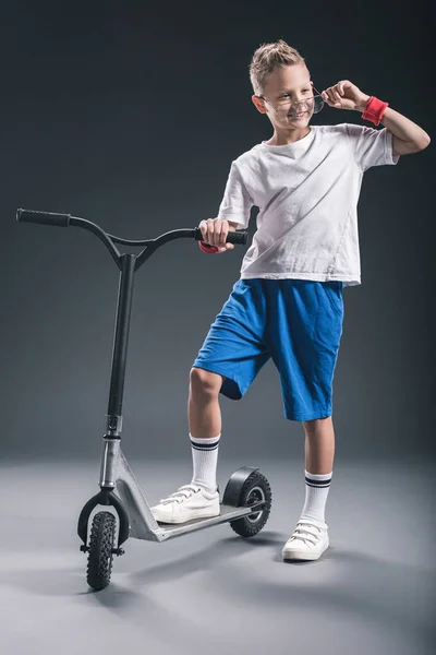 Chico con estilo en gafas con scooter mirando hacia otro lado sobre fondo gris - foto de stock