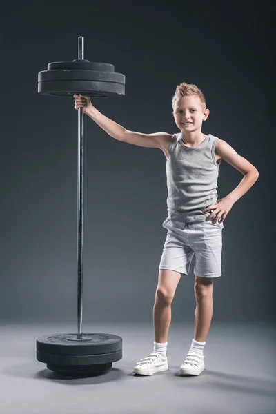 Souriant pré-adolescent garçon en vêtements de sport debout près de Barbell sur fond sombre — Photo de stock