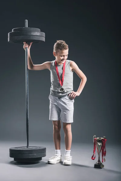 Niño sonriente en ropa deportiva con barra y medalla mirando a la copa de campeones en el fondo oscuro - foto de stock