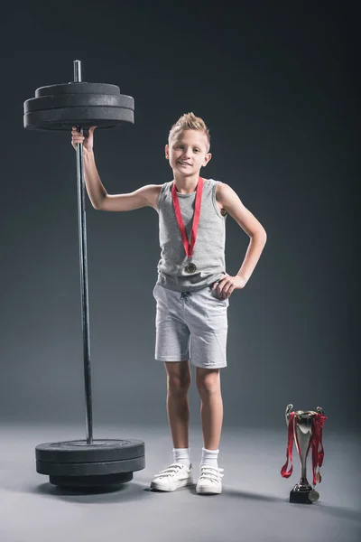 Niño preadolescente en ropa deportiva con barra, copa de campeones y medallas en el fondo oscuro - foto de stock
