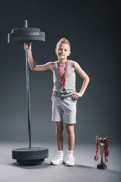 Pre-adolescent boy in sportswear with barbell, champions cup and medals on dark backdrop — Stock Photo