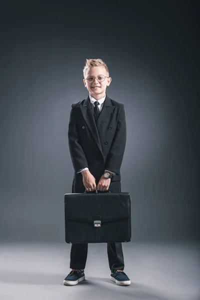 Stylish boy in businessman suit and eyeglasses with suitcase on dark background — Stock Photo