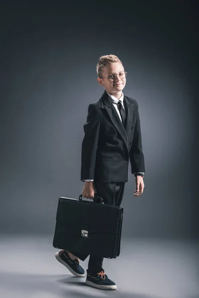 Menino sorridente em terno de homem de negócios e óculos com mala no fundo escuro — Fotografia de Stock