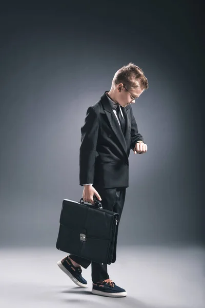 Préadolescent garçon en costume d'homme d'affaires et lunettes avec valise vérifier l'heure sur fond sombre — Photo de stock