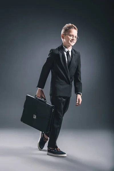 Menino pré-adolescente em terno de homem de negócios e óculos com mala andando sobre fundo escuro — Fotografia de Stock