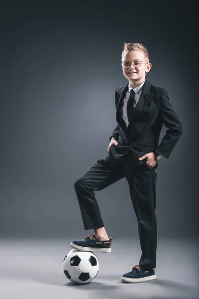 Lächelnder Junge, der als Geschäftsmann mit Fußballball gekleidet in die Kamera auf grauem Hintergrund blickt — Stockfoto