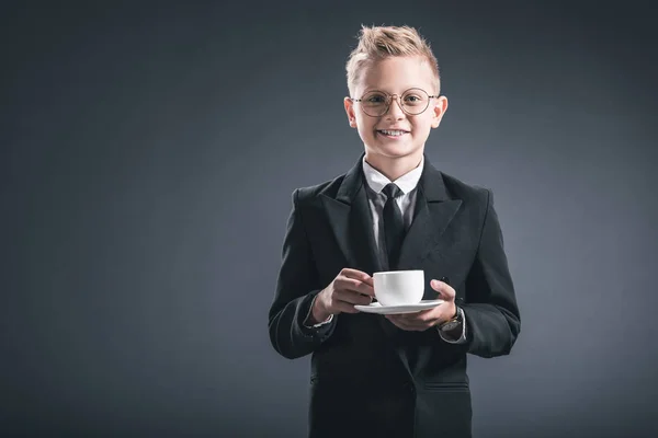 Ritratto di ragazzo sorridente in abito da uomo d'affari e occhiali che tengono la tazza di caffè su sfondo scuro — Foto stock