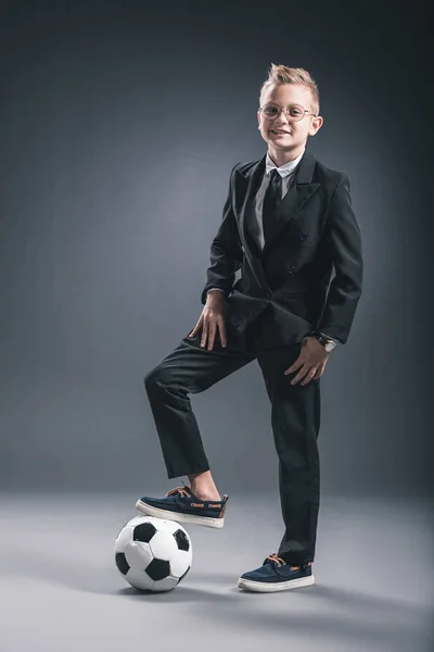 Boy dressed as businessman with soccer ball looking at camera on grey background — Stock Photo