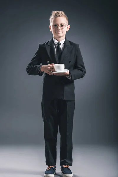 Boy in businessman suit and eyeglasses holding cup of coffee on dark backdrop — Stock Photo