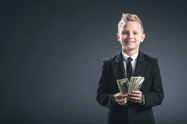 Retrato del niño preadolescente sonriente vestido de hombre de negocios sosteniendo billetes de dólar en manos sobre fondo gris - foto de stock