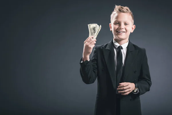Retrato del niño preadolescente sonriente vestido de hombre de negocios sosteniendo billetes de dólar en manos sobre fondo gris - foto de stock