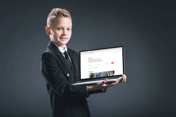 Усміхаючись хлопчика в костюм бізнесмена показ ноутбук з airbnb сайт екран на сірий фон — стокове фото