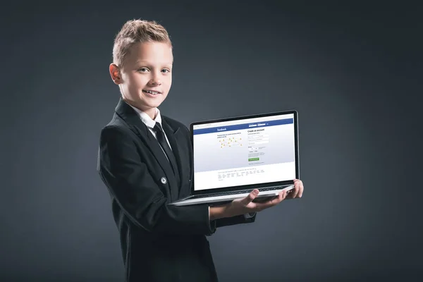 Улыбающийся мальчик в костюме бизнесмена показывает ноутбук с вэб-сайтом facebook на сером фоне — стоковое фото