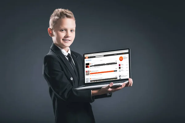 Улыбающийся мальчик в костюме бизнесмена показывает ноутбук с сайтом soundcloud на сером фоне — стоковое фото