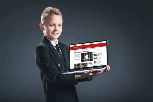 Улыбающийся мальчик в костюме бизнесмена показывает ноутбук с сайтом bbC на сером фоне — стоковое фото