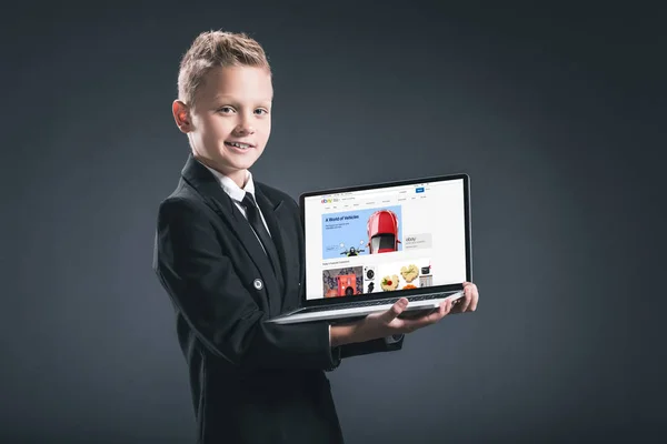 Niño sonriente en traje de hombre de negocios que muestra el ordenador portátil con el sitio web de ebay sobre fondo gris - foto de stock