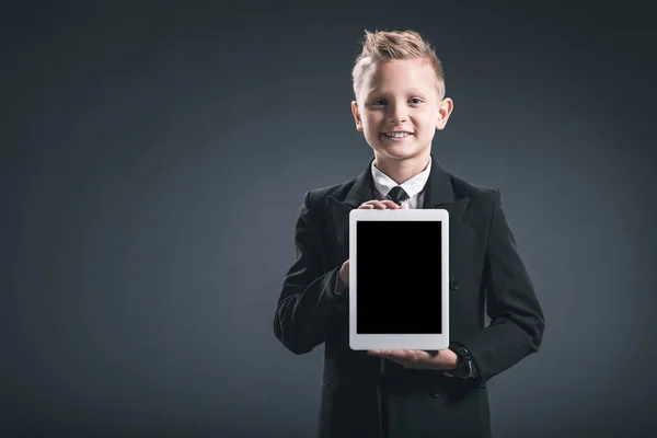 Porträt eines lächelnden Jungen, der wie ein Geschäftsmann gekleidet ist und Tablet mit leerem Bildschirm in der Hand vor grauem Hintergrund zeigt — Stockfoto