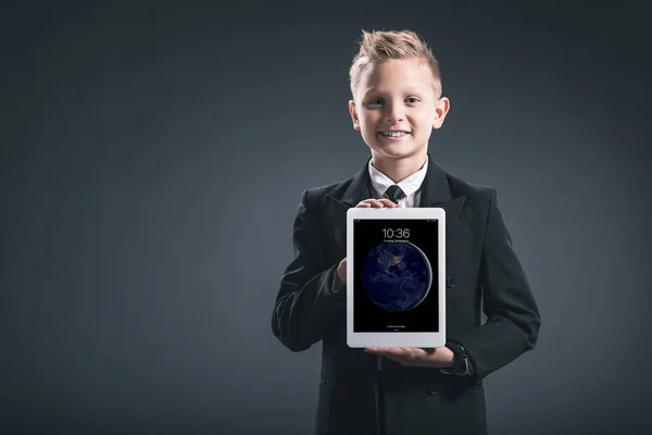 Portrait of smiling boy dressed like businessman showing tablet in hands on grey backdrop — Stock Photo
