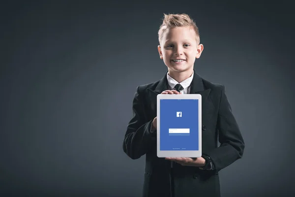Портрет улыбающегося мальчика, одетого как бизнесмен, показывающего планшет с логотипом Facebook в руках на сером фоне — стоковое фото