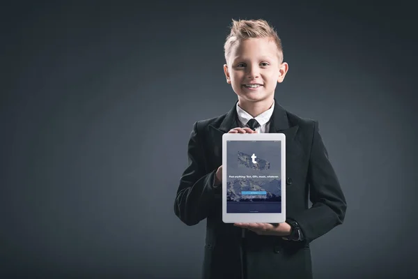 Retrato de niño sonriente vestido como hombre de negocios mostrando la tableta con el logotipo de tumblr en las manos sobre fondo gris - foto de stock