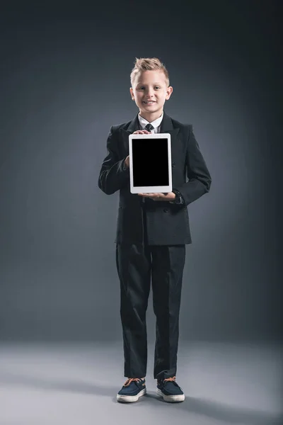 Sorrindo menino vestido como empresário mostrando tablet com tela em branco nas mãos em fundo cinza — Fotografia de Stock