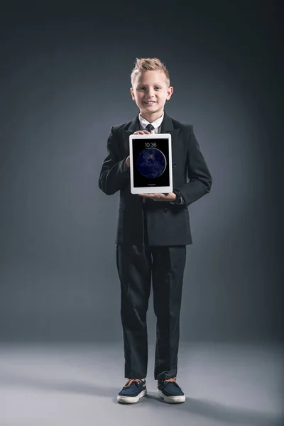 Улыбающийся мальчик, одетый как бизнесмен, показывающий планшет в руках на сером фоне — стоковое фото
