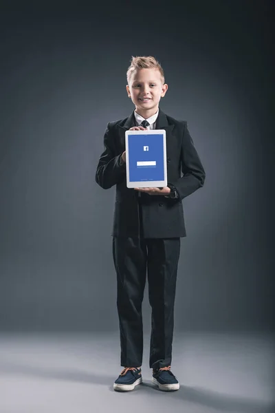 Улыбающийся мальчик, одетый как бизнесмен, показывающий планшет с логотипом Facebook в руках на сером фоне — стоковое фото