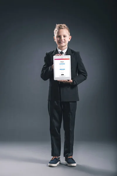 Niño sonriente vestido como hombre de negocios mostrando tableta con el logotipo de instagram en las manos sobre fondo gris - foto de stock