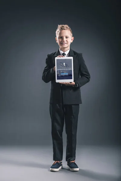 Улыбающийся мальчик, одетый как бизнесмен, показывает планшет с логотипом tumblr в руках на сером фоне — стоковое фото