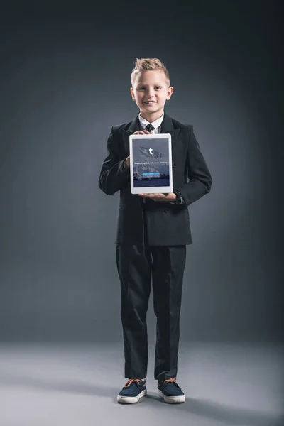 Muchacho sonriente vestido como hombre de negocios mostrando tableta con logotipo tumblr en las manos sobre fondo gris - foto de stock