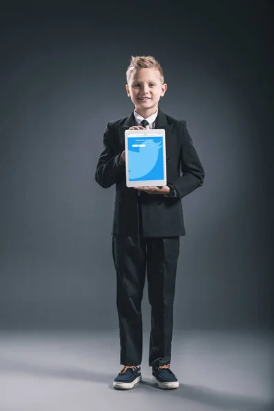 Smiling boy dressed like businessman showing tablet with twitter website in hands on grey backdrop — Stock Photo