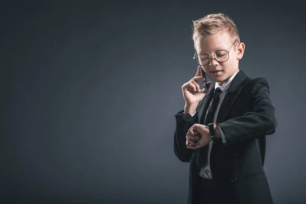 Als Geschäftsmann verkleideter preußischer Junge checkt die Uhrzeit, während er auf dunklem Hintergrund mit dem Smartphone spricht — Stockfoto
