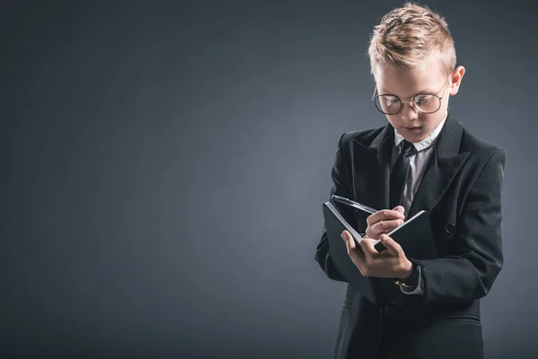 Портрет мальчика в костюме бизнесмена и очках, делающего заметки в блокноте на сером фоне — стоковое фото