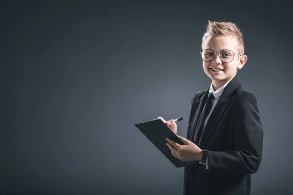 Niño preadolescente sonriente en traje de hombre de negocios y gafas con bloc de notas sobre fondo gris - foto de stock