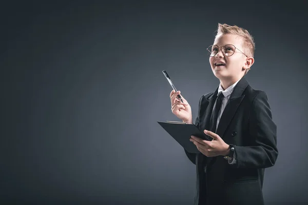 Preadolescente pensativo en traje de hombre de negocios y gafas con bloc de notas sobre fondo gris - foto de stock