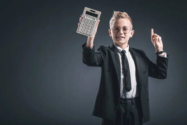 Portrait de garçon habillé en homme d'affaires en lunettes geste et montrant calculatrice sur fond sombre — Photo de stock
