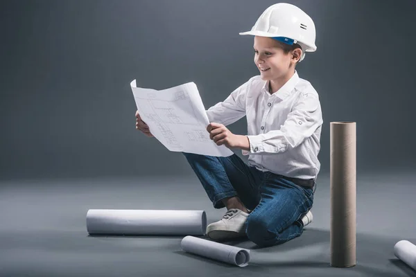 Smiling boy in architect helmet looking at blueprints on dark background — Stock Photo